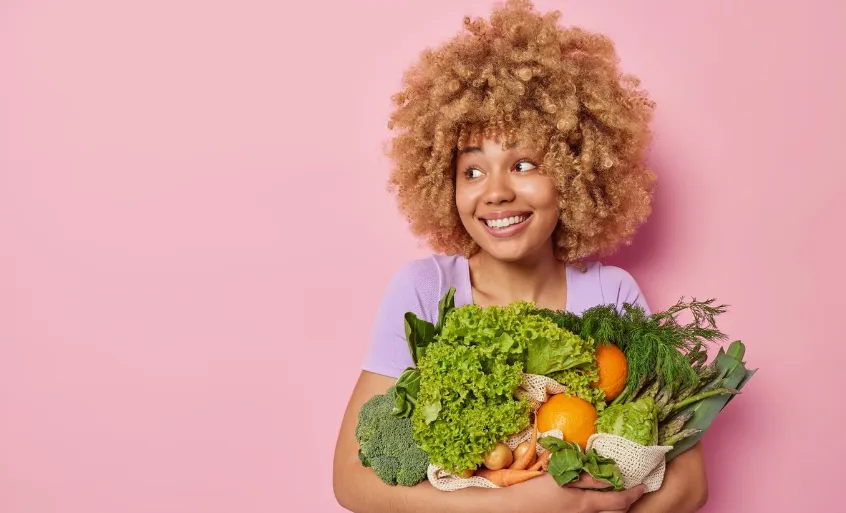 Mulher sorridente segurando uma variedade de vegetais frescos, simbolizando hábitos alimentares saudáveis.