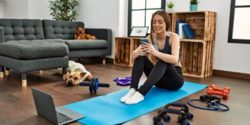 Mulher sorridente em casa, com acessórios de treino como halteres, bandas elásticas e computador, representando a prática de exercícios em casa.