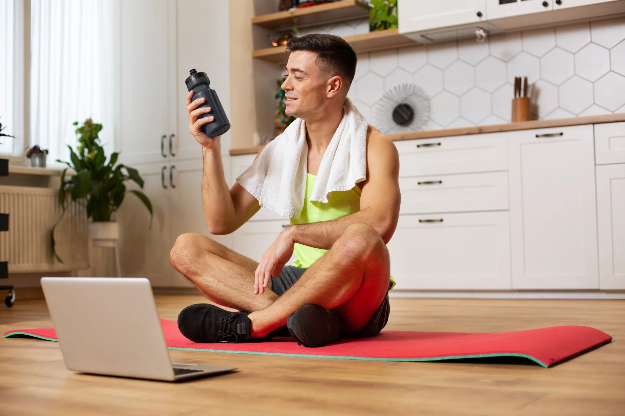 Homem se preparando para o treino aeróbico em casa, sentado em um tapete com uma garrafa de água e laptop à frente.