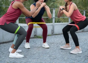 Três mulheres realizando agachamentos com mini bands em um treino ao ar livre.