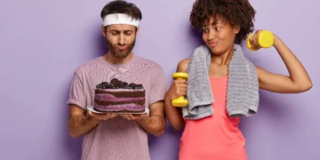 Homem segurando um bolo e mulher levantando um haltere, representando a escolha entre alimentação e treino.