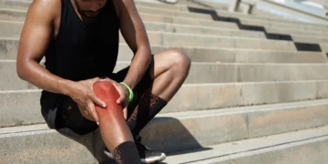 Homem com dor no joelho, sentado em uma escada, segurando o joelho lesionado.