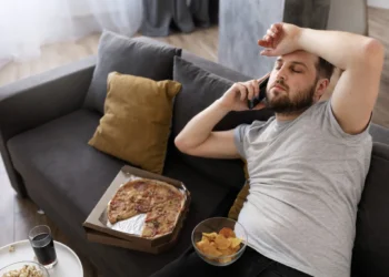 Homem relaxando no sofá comendo fast food, representando o estilo de vida sedentário e a importância de combater esse hábito para melhorar a saúde.