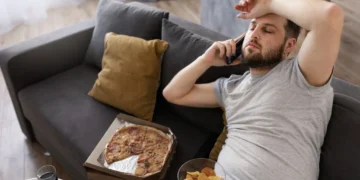 Homem relaxando no sofá comendo fast food, representando o estilo de vida sedentário e a importância de combater esse hábito para melhorar a saúde.