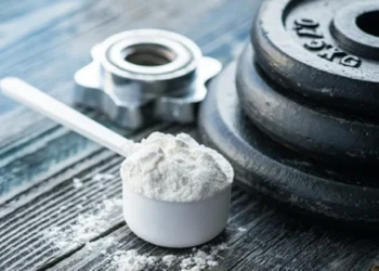 Dose de creatina em pó ao lado de um peso de academia sobre superfície de madeira.