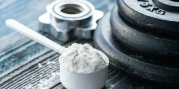 Dose de creatina em pó ao lado de um peso de academia sobre superfície de madeira.