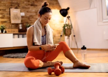 Mulher praticando atividade física em casa, sentada no tapete, descansando enquanto olha para o celular