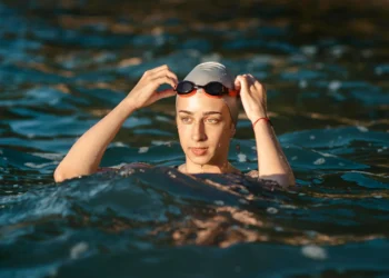 "Mulher nadando ao ar livre com capô de natação e óculos, ajustando os óculos na água."