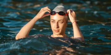"Mulher nadando ao ar livre com capô de natação e óculos, ajustando os óculos na água."