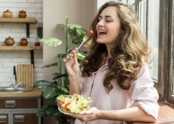 "Mulher sorridente comendo salada fresca em uma cozinha iluminada."