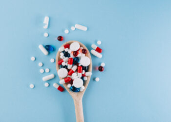 Colher de madeira cheia de comprimidos e cápsulas de diferentes cores espalhados sobre uma superfície azul, simbolizando o uso indiscriminado de medicamentos.