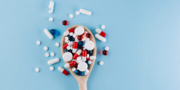 Colher de madeira cheia de comprimidos e cápsulas de diferentes cores espalhados sobre uma superfície azul, simbolizando o uso indiscriminado de medicamentos.