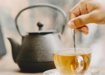 Uma mão mexendo chá quente em uma xícara, com uma chaleira ao fundo, sobre uma mesa de madeira