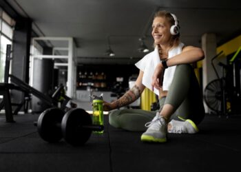 Mulher se exercitando em uma academia, fazendo pausa com fones de ouvido e garrafa de água ao lado de pesos.