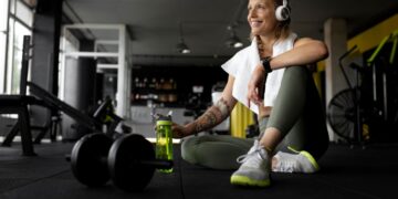 Mulher se exercitando em uma academia, fazendo pausa com fones de ouvido e garrafa de água ao lado de pesos.