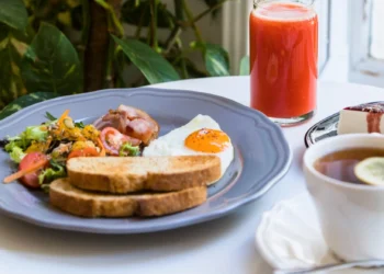 Café da manhã com ovos, salada fresca, bacon, torradas, suco vermelho e chá, servido sobre uma mesa branca.