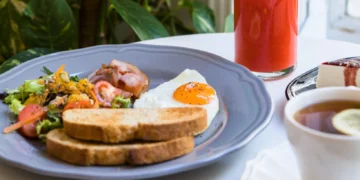 Café da manhã com ovos, salada fresca, bacon, torradas, suco vermelho e chá, servido sobre uma mesa branca.