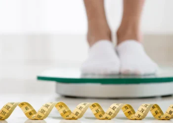 Pessoa pesando-se em uma balança digital com fita métrica ao lado.