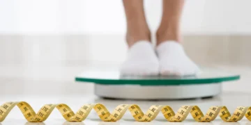 Pessoa pesando-se em uma balança digital com fita métrica ao lado.