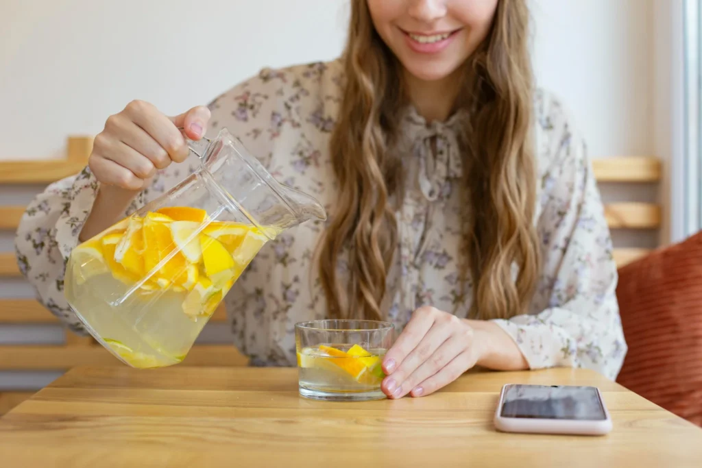 Pessoa preparando água com limão, colocando pedaços de laranja em um copo, ilustrando o modo de consumo da bebida.