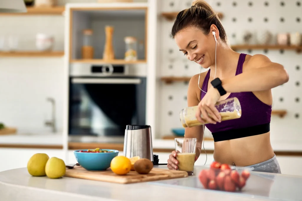 Mulher sorridente em roupa esportiva preparando smoothie de frutas na cozinha pela manhã.