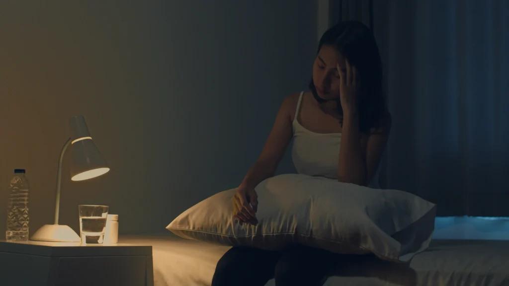 Mulher jovem asiática sentada na cama tomando um remédio para insônia, representando os efeitos do transtorno no sono.