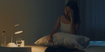 Mulher jovem asiática sentada na cama tomando um remédio para insônia, representando os efeitos do transtorno no sono.