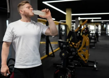 Homem bebendo creatina em garrafa plástica durante treino de musculação na academia.