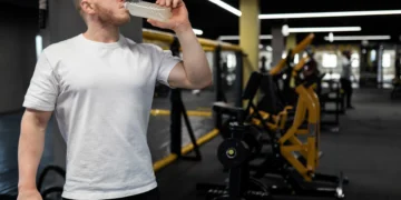 Homem bebendo creatina em garrafa plástica durante treino de musculação na academia.