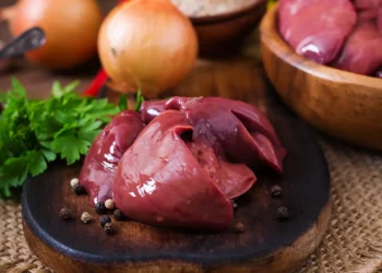 Fígado de boi cru com temperos, cebolas e ervas frescas sobre uma superfície de madeira.