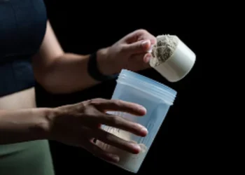 Pessoa segurando um copo e medidor de whey protein para preparar suplemento.