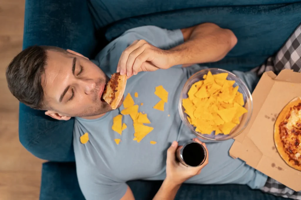 Homem deitado no sofá consumindo alimentos ultraprocessados, como pizza e salgadinhos.