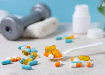 Suplementos e cápsulas variadas em colher e espalhadas na mesa, com um haltere desfocado ao fundo.
