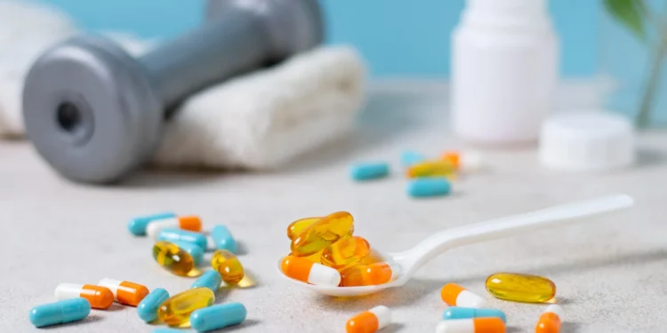 Suplementos e cápsulas variadas em colher e espalhadas na mesa, com um haltere desfocado ao fundo.