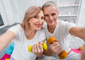 Casal maduro sorridente fazendo exercícios com halteres em casa, promovendo saúde e autoestima após os 40 anos.