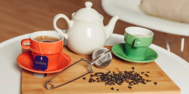Xícara de chá Earl Grey ao lado de uma cafeteira e folhas de chá em uma tábua de madeira, representando a escolha entre café e chá.