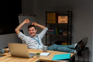 Homem sorrindo e relaxando com os pés na mesa, mostrando satisfação e descanso no ambiente de trabalho.