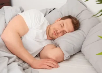 Homem dormindo confortavelmente no lado esquerdo da cama, com expressão tranquila e coberto por lençóis cinza.