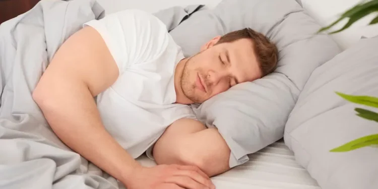 Homem dormindo confortavelmente no lado esquerdo da cama, com expressão tranquila e coberto por lençóis cinza.