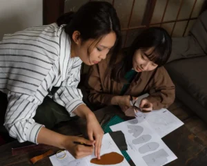 Duas mulheres colaborando em uma oficina de design, uma desenhando e a outra cortando materiais, simbolizando o trabalho em equipe no aprendizado.