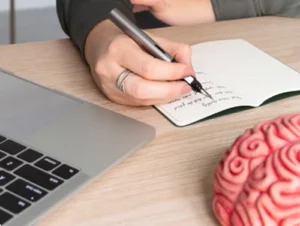 Pessoa escrevendo em caderno próximo a um modelo de cérebro e laptop, simbolizando o cuidado com a memória.