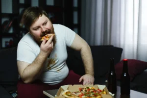 Homem comendo pizza enquanto está sentado no sofá, com garrafas de cerveja ao lado, refletindo hábitos alimentares desatentos.