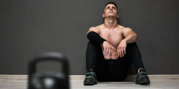 Homem cansado sentado no chão após treino intenso na academia, refletindo sobre o tempo ideal de descanso entre as séries.
