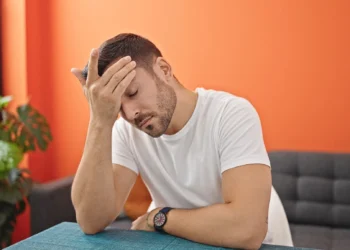 Homem cansado com a mão na testa, demonstrando exaustão mental sentado à mesa em uma sala de jantar.