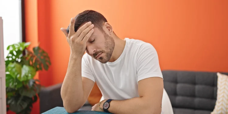 Homem cansado com a mão na testa, demonstrando exaustão mental sentado à mesa em uma sala de jantar.