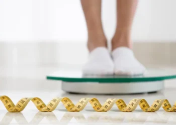 Pessoa em pé na balança digital com fita métrica ao fundo, ilustrando o melhor horário para medir o peso real.