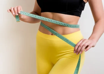 Mulher medindo a cintura com fita métrica, usando roupas esportivas amarelas e preta.