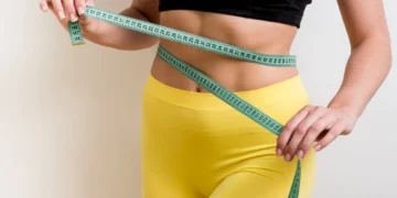 Mulher medindo a cintura com fita métrica, usando roupas esportivas amarelas e preta.