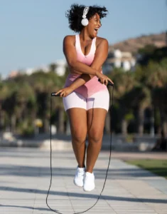 Mulher feliz pulando corda ao ar livre, usando roupa esportiva rosa e fones de ouvido, demonstrando energia e bem-estar.