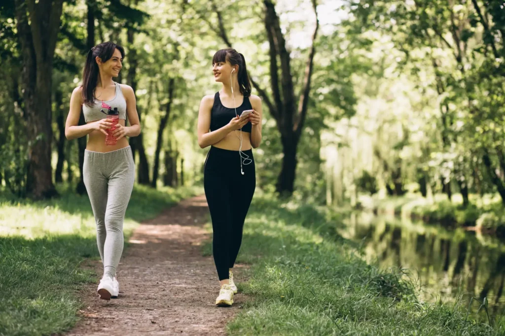 Duas mulheres caminhando em um parque verde, sorrindo e conversando durante uma caminhada ao ar livre.